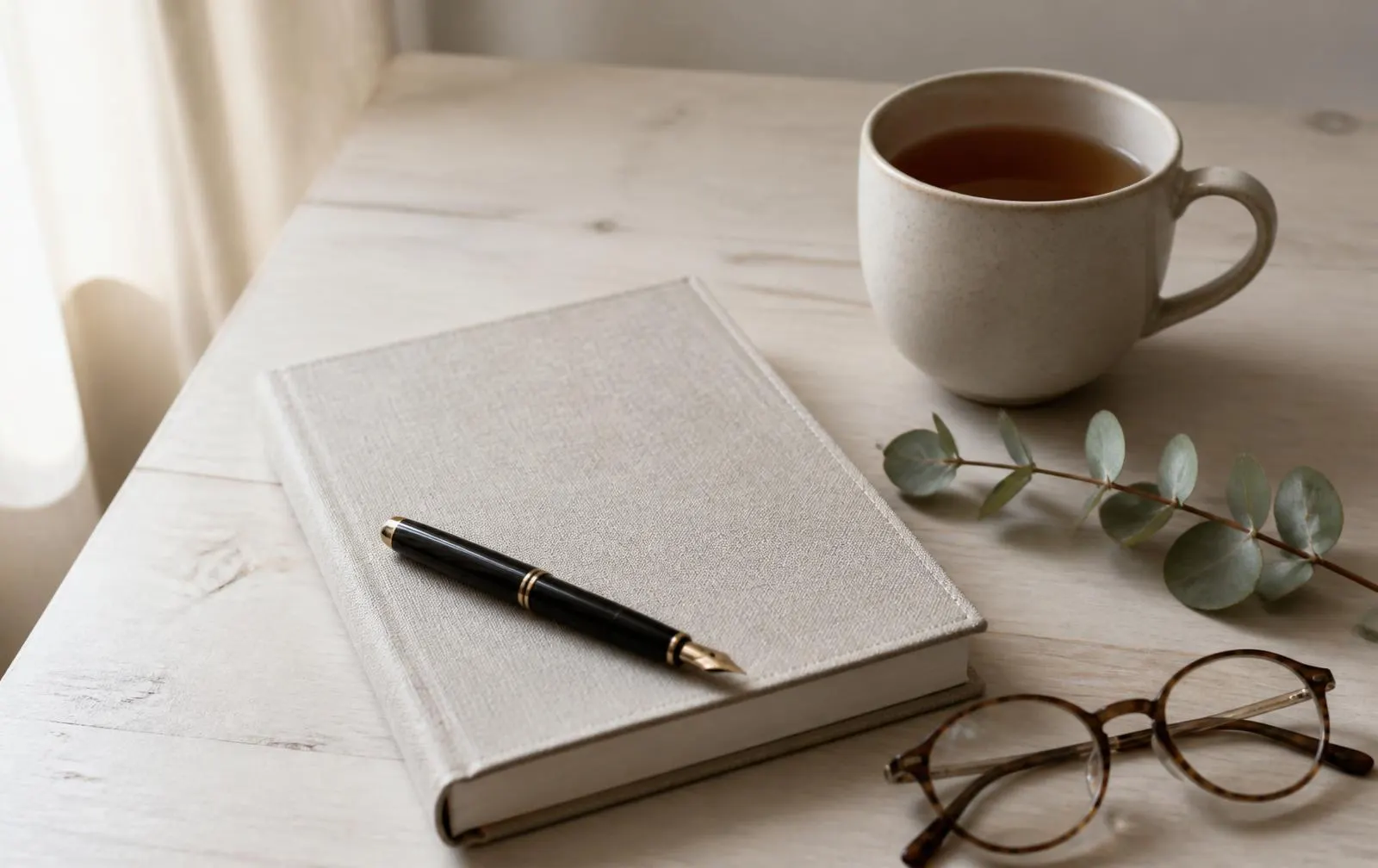 A linen notebook, fountain pen, reading glasses, tea, and a eucalyptus sprig on a pale wooden table